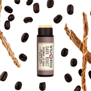 Dark Roast Lip Balm in a biodegradable paper tube. Cap is removed revealing a creamy lip balm. Coffee beans and vetiver root surround.