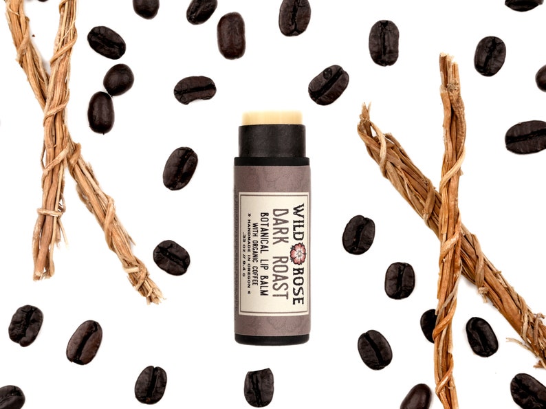 Dark Roast Lip Balm in a biodegradable paper tube. Cap is removed revealing a creamy lip balm. Coffee beans and vetiver root surround.