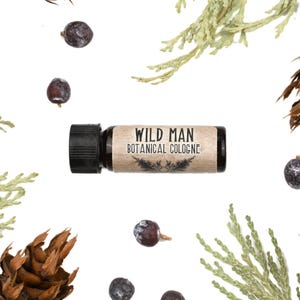 May include: A small cylindrical bottle of Wild Man Botanical Cologne with a black cap and a tan label featuring black text and a small botanical illustration, surrounded by scattered dark purple juniper berries and green cedar sprigs.