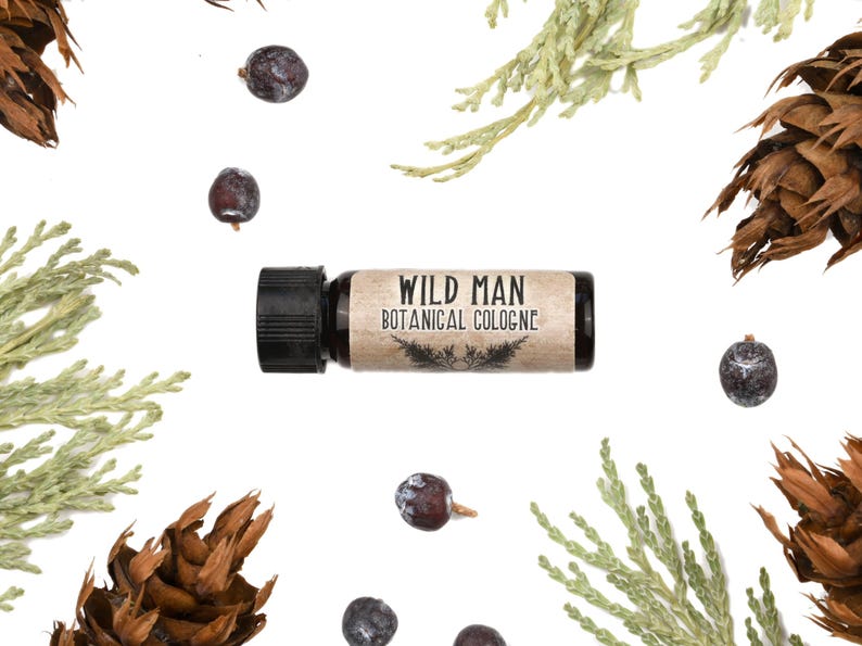 May include: A small cylindrical bottle of Wild Man Botanical Cologne with a black cap and a tan label featuring black text and a small botanical illustration, surrounded by scattered dark purple juniper berries and green cedar sprigs.