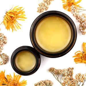 Renewal Healing Salve in a amber glass jars. Lids are removed revealing a creamy yellow balm. Dried yarrow and calendula surround.