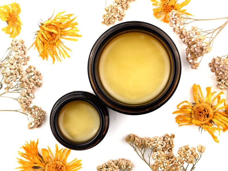 Renewal Healing Salve in a amber glass jars. Lids are removed revealing a creamy yellow balm. Dried yarrow and calendula surround.