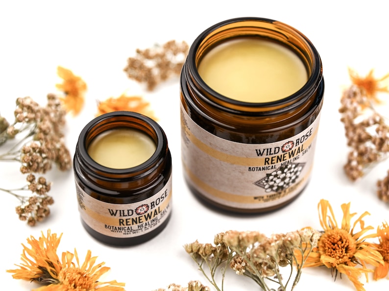 Renewal Healing Salve in a amber glass jars. Lids are removed revealing a creamy yellow balm. Dried yarrow and calendula surround.