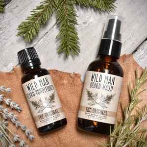 May include: Two amber glass bottles with black caps and labels. The bottles contain beard wash and beard conditioner. The labels are white with black text and a small illustration of a tree. The text on the labels reads "Wild Man Beard Wash" and "Wild Man Beard Conditioner". The text on the labels also reads "The Original" and "With Cedar & Lime".