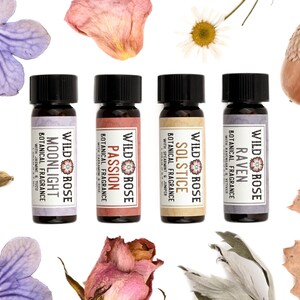 May include: Four small glass bottles with black caps, each featuring a label reading Wild Rose Botanical Fragrance with specific scent names: Moonlight, Passion, Solstice, and Raven. The bottles are surrounded by purple flowers, dried rose petals, a daisy, an acorn, cardamom pods, and a dried oak leaf.