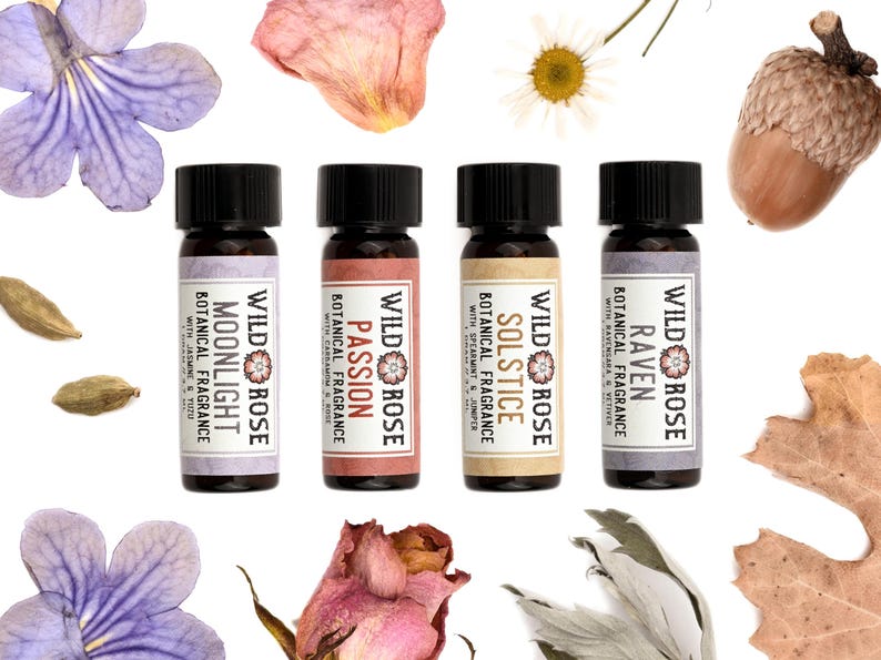 May include: Four small glass bottles with black caps, each featuring a label reading Wild Rose Botanical Fragrance with specific scent names: Moonlight, Passion, Solstice, and Raven. The bottles are surrounded by purple flowers, dried rose petals, a daisy, an acorn, cardamom pods, and a dried oak leaf.
