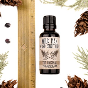 Wild Man Beard Conditioner in The Original scent shown in a 30ml amber glass bottle. Bottle is next to a ruler measuring about 3 3/4" tall. Cedar, fir cones and juniper berries surround.