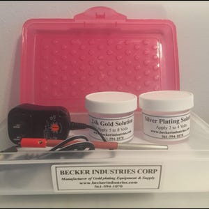 May include: A clear plastic storage box with a pink lid containing a black power adapter, a red and silver plating wand, and two white jars labeled 24k Gold Solution and Silver Plating Solution. A label reads Becker Industries Corp.