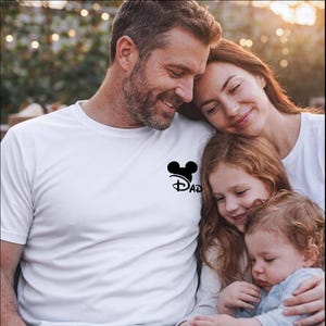 May include: A white short-sleeved t-shirt featuring a black graphic on the left chest area. The graphic consists of a silhouette of mouse ears positioned above the word DAD, written in a stylized, cursive font.