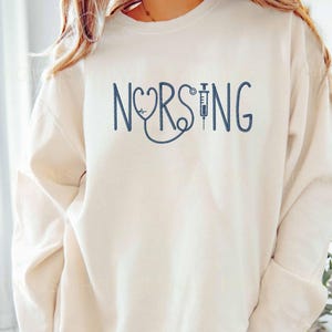 May include: Cream-colored sweatshirt with the word "NURSING" in navy blue script. The "O" in "NURSING" is replaced with a stethoscope and heart symbol, and the "I" is a syringe. A casual top for healthcare professionals.
