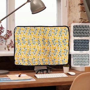 以下が含まれることがあります： 黄色い花柄、黒いトリム、お揃いの柄のディスプレイスタンドが付いたコンピューターモニターカバー。カバーは、白い背景に黄色の花と緑の茎の繰り返しデザインが特徴です。スタンドには3つの異なる生地パターンが表示されます。