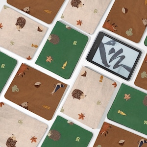 May include: An assortment of fabric e-reader covers in various colors, including brown, green, and beige. Each cover features embroidered woodland-themed designs such as hedgehogs, foxes, and leaves. An e-reader with a black screen is also visible.