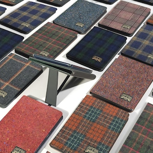 May include: A collection of rectangular tablet cases featuring various wool-like tweed and plaid fabric patterns in colors including blue, green, red, and brown. Each case has a small rectangular metal logo plate and a black folding stand.