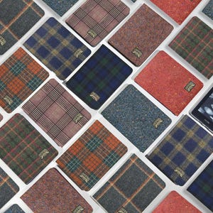 Könnte beinhalten: Eine Sammlung rechteckiger Schutzhüllen für E-Reader mit verschiedenen Tweed- und Karomustern in Farben wie Rot, Blau, Grün, Orange und Grau. Jede Hülle ist mit einem kleinen, rechteckigen Metallschild mit Markenlogo versehen.