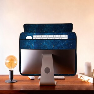 Può includere: Una copertura per monitor di computer blu scuro con un motivo celeste di stelle dorate e lune crescenti. Sono visibili una tastiera e un mouse bianchi. La copertura è posizionata su un monitor di computer argentato su una scrivania di legno.