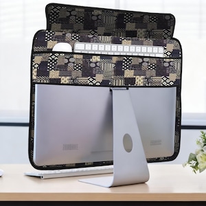 Puede incluir: Una funda para monitor de ordenador con estampado negro y marrón con bolsillos para teclado y ratón. La funda está sobre un monitor de ordenador plateado, con un teclado y un ratón blancos sobre el escritorio. Un pequeño ramo de flores blancas está a la derecha.
