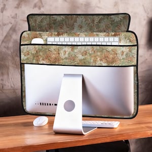 Puede incluir: Un monitor de computadora con una cubierta con estampado floral. La cubierta es verde, marrón y beige. Se ven un teclado y un ratón blancos. El monitor está sobre un escritorio de madera.