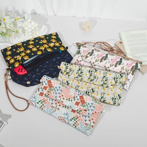Op de afbeelding: Een assortiment stoffen pouches met bloempatronen. De pouches hebben verschillende ontwerpen, waaronder gele madeliefjes, roze en rode bloemen en groene bladeren. Eén pouch is open en onthult een zwart interieur en een rood accessoire.