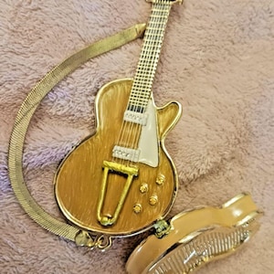 May include: A gold-toned guitar-shaped pendant necklace with a matching bracelet. The guitar pendant features a detailed design with a white pickguard and gold hardware. The bracelet is a hinged style with a textured surface. The jewelry is set against a pink background.