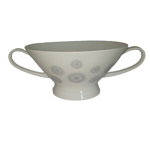 May include: A white porcelain bowl featuring two curved handles and a pedestal base. The exterior surface is decorated with a repeating pattern of small, light gray circular starburst or floral motifs arranged in a symmetrical layout.