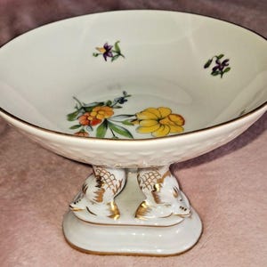May include: A white porcelain pedestal bowl with a gold rim and floral designs. The bowl features a textured exterior and is supported by a base with two decorative, gold-accented figures. The floral design includes yellow, orange, and purple flowers.