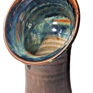 May include: Handmade ceramic vase with a unique design. The vase features a wide, open top with a handle, and a dark brown exterior. The interior is a blend of blue, green, and brown hues, creating a striking contrast. The vase is a decorative piece.