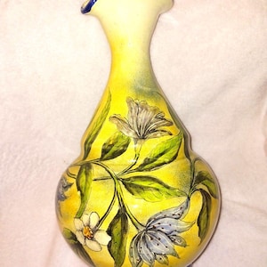 May include: A hand-painted ceramic vase with a yellow background and floral design. The vase features a flared top with a blue rim, a bulbous body, and a blue base. The flowers are white and blue with green leaves.