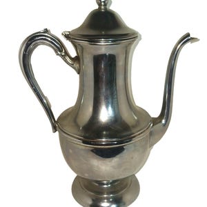 Puede incluir: Una cafetera de metal color plata con base redondeada, cuello alto, asa curva y un pico largo y delgado. La cafetera incluye una tapa extraíble con un pequeño remate puntiagudo en la parte superior y se asienta sobre una base circular.