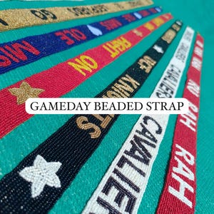May include: A collection of beaded straps in various colors, including red, blue, and gold. Each strap features text and designs like stars and hearts. The text "GAMEDAY BEADED STRAP" is also visible.
