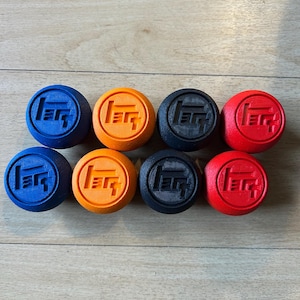 May include: Eight round objects in various colors: blue, orange, black, and red. Each object has a raised, stylized logo in the center. The objects are arranged in two rows of four, against a light brown wooden surface.