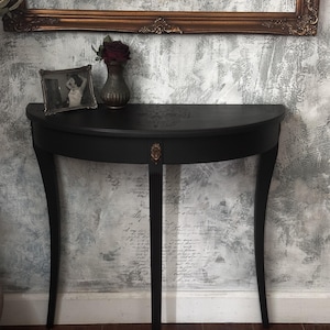May include: A semi-circular black console table with three slender, curved legs and a central decorative bronze-toned accent piece. The tabletop features a subtle, dark skull motif etched into the surface, providing a distinct visual detail.