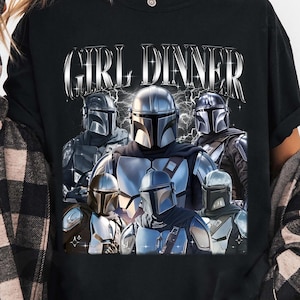 May include: Black t-shirt featuring a graphic of several Mandalorian characters with the text "GIRL DINNER" in a silver, stylized font. The image has a vintage, distressed look. The shirt is a crew neck style.