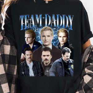 May include: Black t-shirt with the text "TEAM DADDY" in blue and white, featuring images of several men. The shirt has a crew neck and short sleeves.