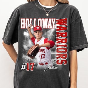 Può includere: Una t-shirt grigio antracite a maniche corte con una stampa grafica di un giocatore di baseball in divisa bianca e rossa con il testo HOLLOWAY WARRIORS, #17 e il nome Jake. Il design include una palla da baseball, un guanto e luci.