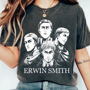 May include: Dark gray t-shirt featuring a black and white illustration of four anime characters. The text "ERWIN SMITH" is printed in white below the illustration. The shirt has a crew neck and short sleeves.