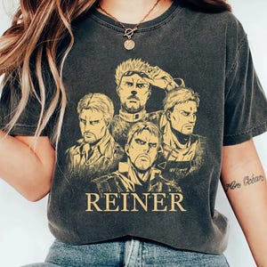 May include: Dark gray t-shirt featuring a graphic of four male characters with stern expressions. The word "REINER" is printed in a bold font below the image. The shirt has short sleeves and a crew neck.
