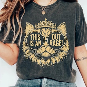 May include: Dark gray t-shirt featuring a gold illustration of a cat wearing sunglasses and a crown. The text on the shirt reads "THIS IS AN OUT RAGE!". The shirt is a casual style.