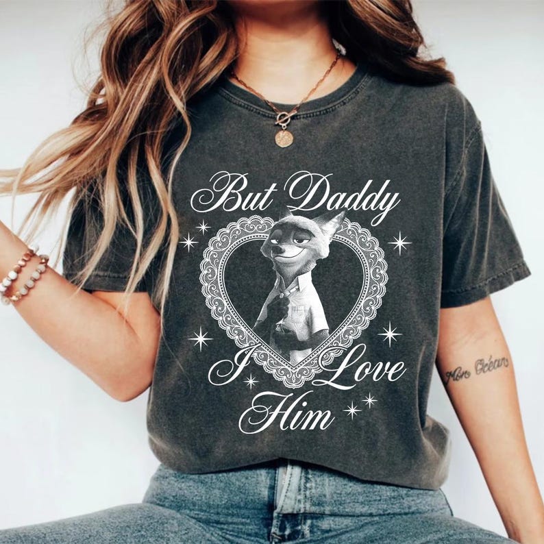 K&ouml;nnte beinhalten: Dunkelgraues T-Shirt mit einer Cartoon-Fuchsfigur in einem herzf&ouml;rmigen Rahmen. Der Text "But Daddy I Love Him" steht oberhalb und unterhalb des Bildes in einer dekorativen Schriftart. Das T-Shirt hat einen Vintage-Look.
