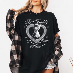 K&ouml;nnte beinhalten: Schwarzes T-Shirt mit einer Cartoon-Fuchsfigur in einem herzf&ouml;rmigen Rahmen. Der Text "But Daddy I Love Him" steht oberhalb und unterhalb des Bildes in einer dekorativen Schriftart. Das T-Shirt ist ein l&auml;ssiger Stil.