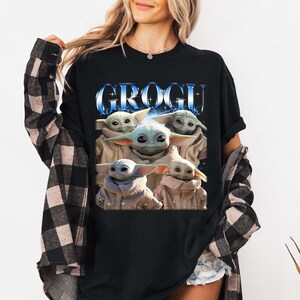 Może przedstawiać: Czarny t-shirt z napisem "GROGU" w niebieskiej, metalicznej czcionce nad grafiką Grogu, znanego r&oacute;wnież jako Baby Yoda, z serii Star Wars. Grafika przedstawia Grogu w beżowym płaszczu.