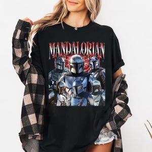 Pu&ograve; includere: T-shirt nera con una grafica di pi&ugrave; personaggi Mandaloriani in armatura argentata e grigia, con la scritta "MANDALORIAN" in testo rosso e bianco. Il design ha un aspetto vintage e usurato.