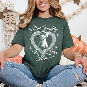 K&ouml;nnte beinhalten: Dunkelgr&uuml;nes T-Shirt mit dem wei&szlig;en Schriftzug "But Daddy I Love Him". Das Shirt hat ein herzf&ouml;rmiges Design mit einer Cartoonfigur. Die Person tr&auml;gt eine blaue Jeans.