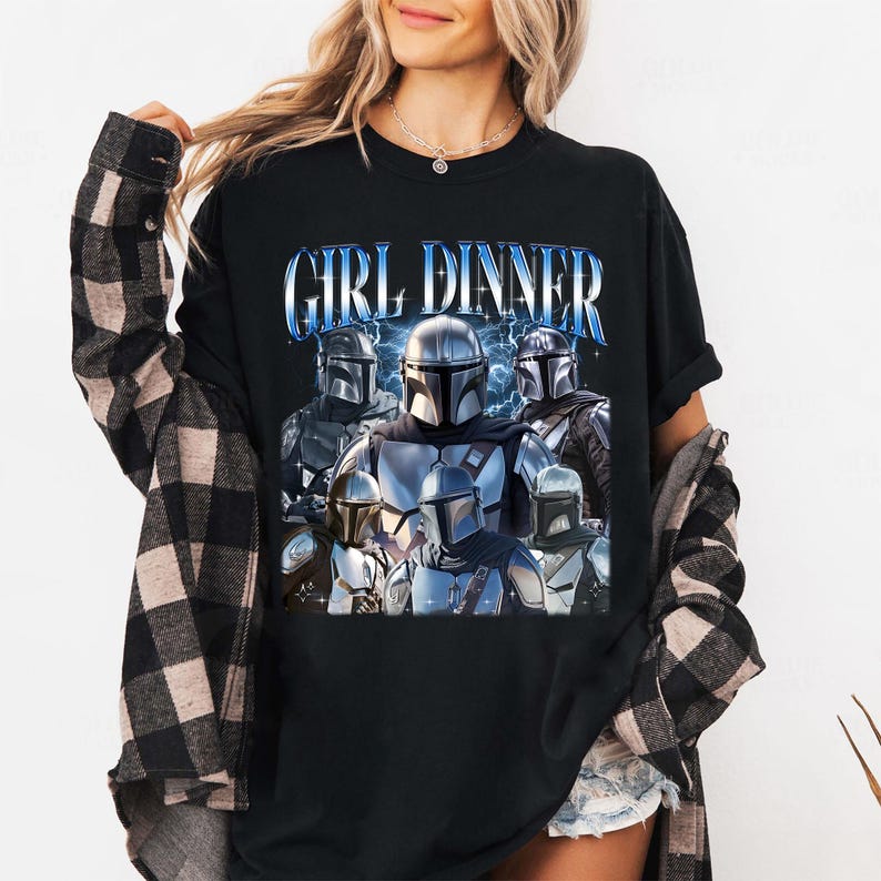 May include: Black t-shirt featuring a graphic of several Mandalorian characters with the text "GIRL DINNER" in blue. The design includes lightning bolts and a futuristic aesthetic. The shirt is worn by a person with long blonde hair.