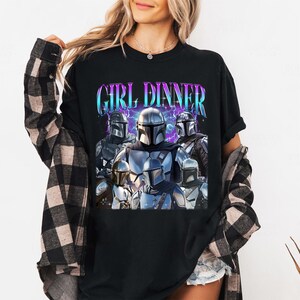 May include: Black t-shirt featuring a graphic of several Mandalorian characters with the text "GIRL DINNER" in a blue and purple font. The design includes lightning bolt accents. The shirt is worn by a person.