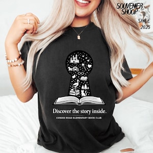 May include: Black t-shirt with a keyhole design showcasing a fairytale scene. The keyhole reveals a castle, carriage, and other magical elements. The text "Discover the story inside" and "COWAN ROAD ELEMENTARY BOOK CLUB" are printed below.