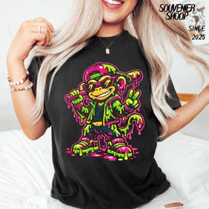 May include: Black t-shirt featuring a cartoon monkey wearing sunglasses, a backwards cap, and a jacket. The monkey is in a vibrant neon green and pink color scheme. The text "SOUVENIER SHOOP SINCE 2025" is printed on the upper right.