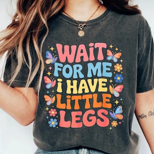 Peut inclure: T-shirt gris foncé avec l'inscription "WAIT FOR ME i HAVE LITTLE LEGS" dans un style rétro coloré. Le motif comprend des papillons et des fleurs roses, bleus, jaunes et oranges.