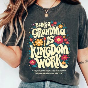 Puede incluir: Camiseta gris oscuro con el texto "Being a Grandma is Kingdom Work" en un estilo retro, rodeada de flores coloridas y pinceles. La camiseta también incluye un versículo bíblico del Salmo 71:18.