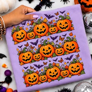 Peut inclure: Un t-shirt violet clair avec un motif horizontal répétitif de citrouilles jack-o'-lantern orange, de feuilles vertes, de chauves-souris violettes, de papillons orange et de bonbons maïs jaunes et blancs imprimés sur le devant.