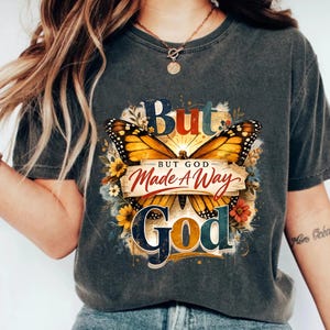 Peut inclure: T-shirt gris foncé avec un motif de papillon coloré et l'inscription "But God Made A Way". Le motif comprend des éléments floraux et une esthétique vintage. Un t-shirt confortable et décontracté.